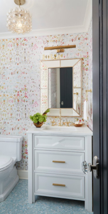 Pink and gold girl's bathroom boasts a beveled mirror mounted on a wall covered in Kandy Brit Pop Wallpaper over a white washstand accented with gold pulls and a white quartz countertop fitted with a polished brass gooseneck faucet illuminated by a brass picture fixed over the mirror and Jamie Young Small Cici 3 Light Drum Pendant hung in the center of the room over blue herringbone floor tiles.