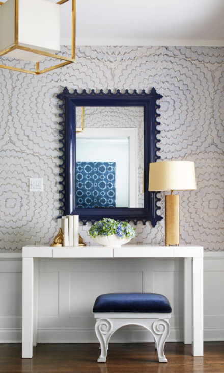 Illuminated by a brass Caged Lantern with Paper Shade a West Elm Parsons Desk with Drawers sits against a wainscoted wall above a blue velvet bench and beneath a navy blue scalloped mirror mounted on gray floral wallpapered walls and illuminated by a gold table lamp complementing gold book ends in this beautiful navy blue and gold foyer.