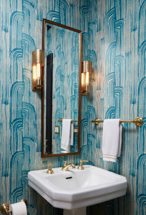 Brightly colored Kelly Wearstler Crescent Wallpaper covers the walls of this stunning powder room featuring a brass vanity mirror flanked by brass sconces mounted above a white pedestal sink finished with a polished brass vintage faucet kit complementing the brass towel bar fixed on an adjacent wall.
